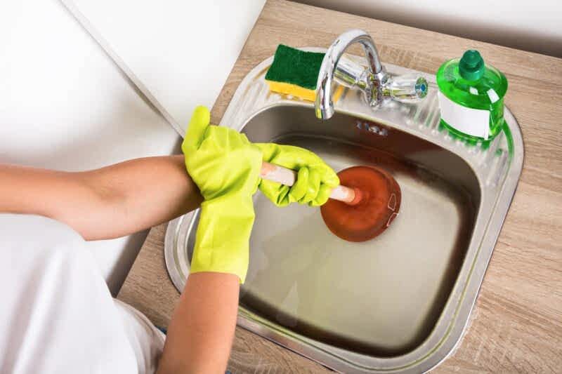 houston homeowner wearing rubber gloves while using a plunger in their kitchen sink full of water