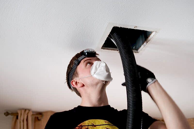 friendly ac technician wearing N95 face mask and headlamp while feeding a large black tube into a living room air vent during duct cleaning